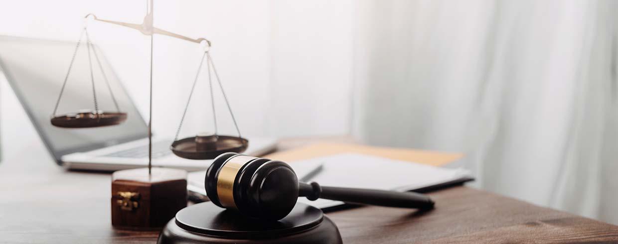Image of a laptop, legal scales and a gavel on a table in front of a bright light window.